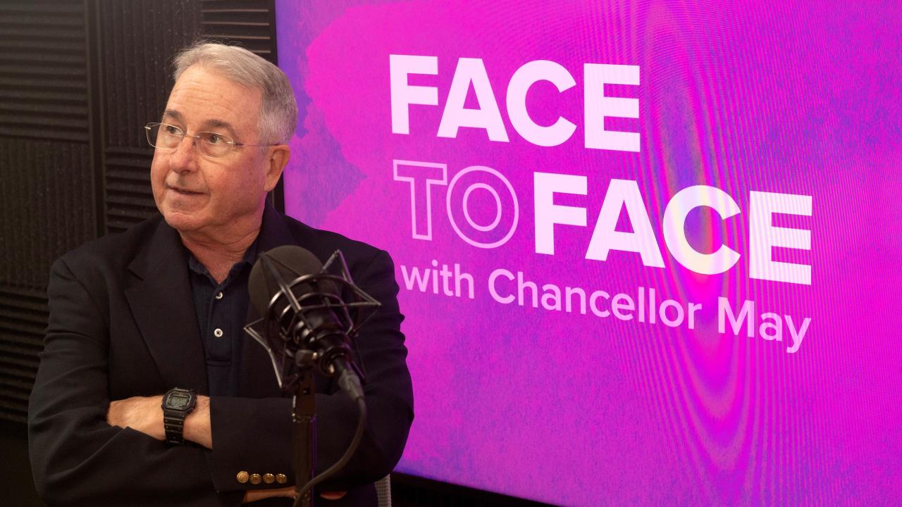 Inside a recording studio, in front of a purple backdrop with the logo for Face to Face with Chancellor Gary S. May sits a man with his arms crossed wearing glasses and short grey hair, staring to their right. 