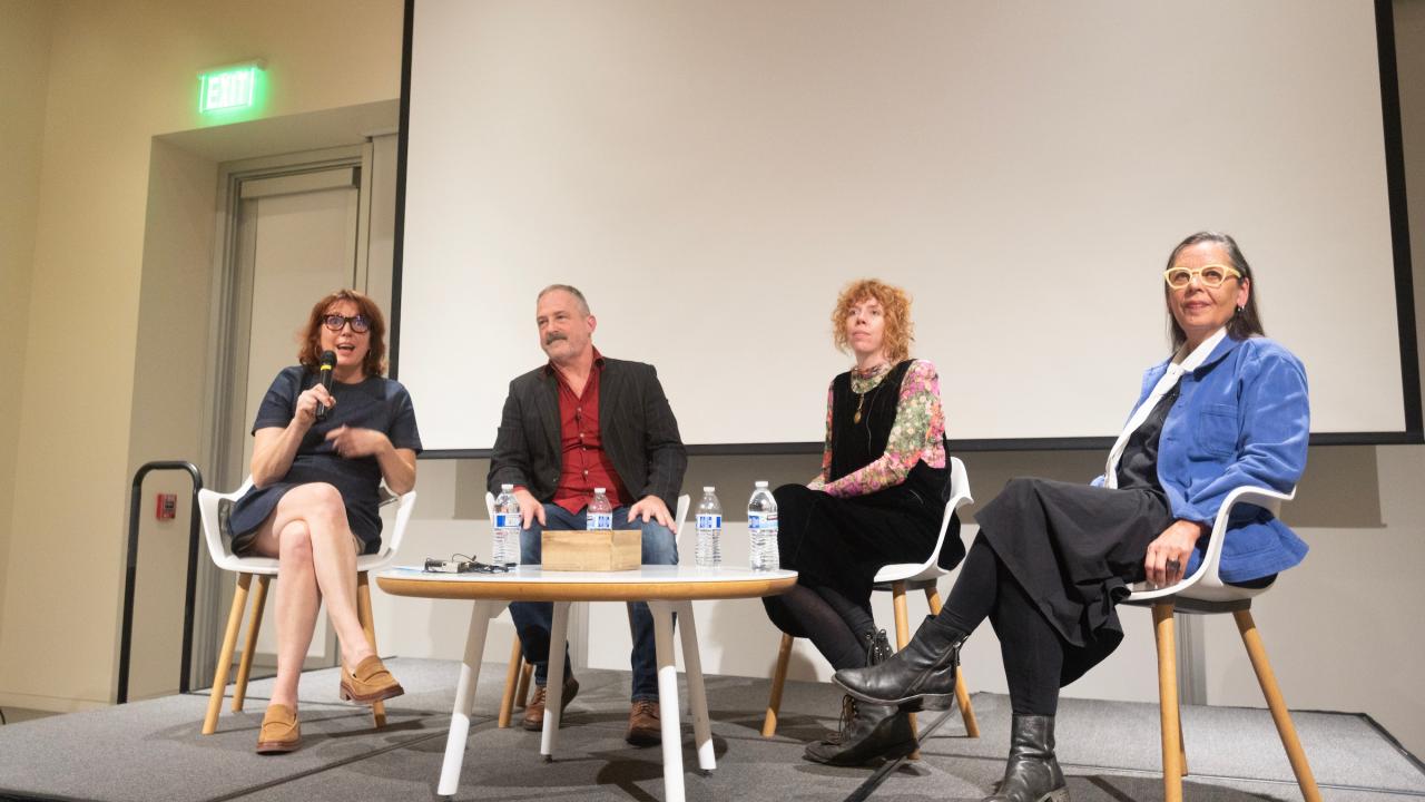 four individuals are seated on top of a small stage in front of a projector screen with a small circular table in front of them