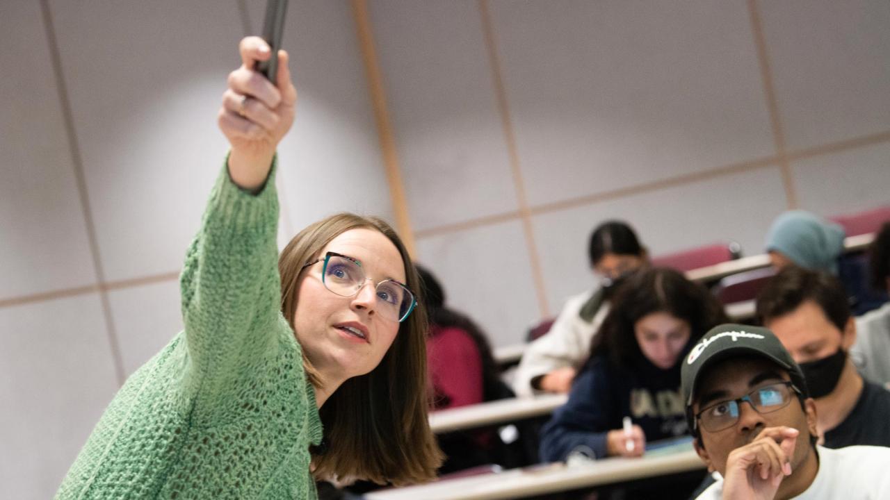 A woman professor points up to the board while students look on