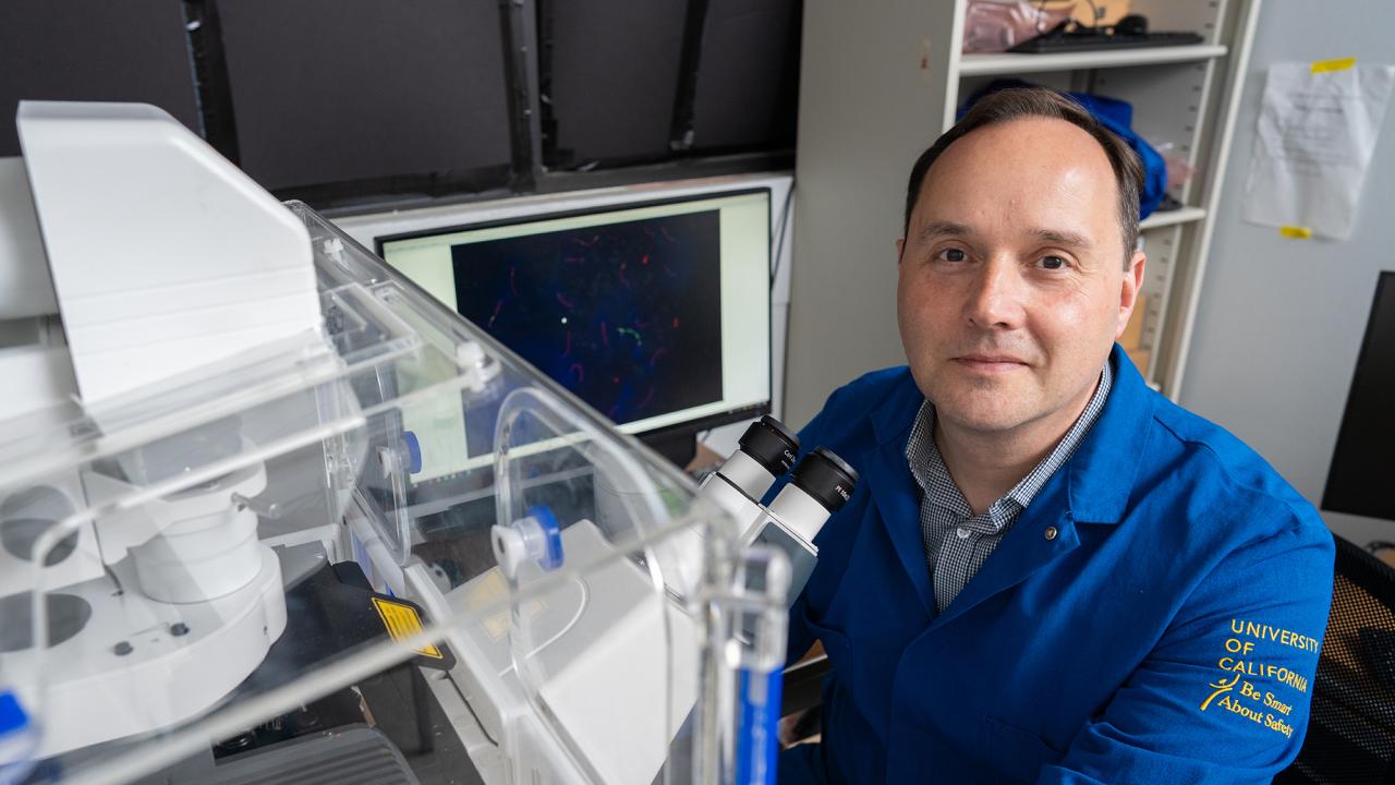 A man in a dark blue labcoat sits in front of a large microscope looking up towards the camera. 