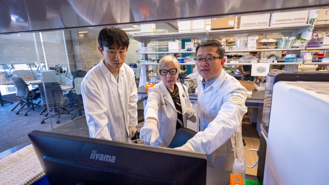 In a laboratory environment, three people gather around a computer monitor, with two of them pointing at the screen.