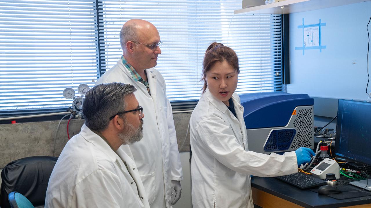 A seated male with grey hair and glasses, a standing bald male with glasses and a younger female with long brown hair tied in a ponytail stand in front of lab equipment. All three wear white lab coats and gloves. 
