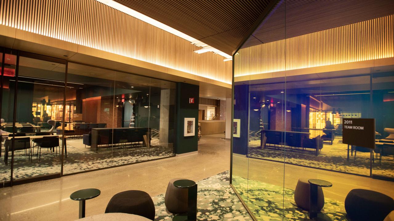 An angular view of two brightly lit, glass offices converging in a carpeted common space with chairs and a table.