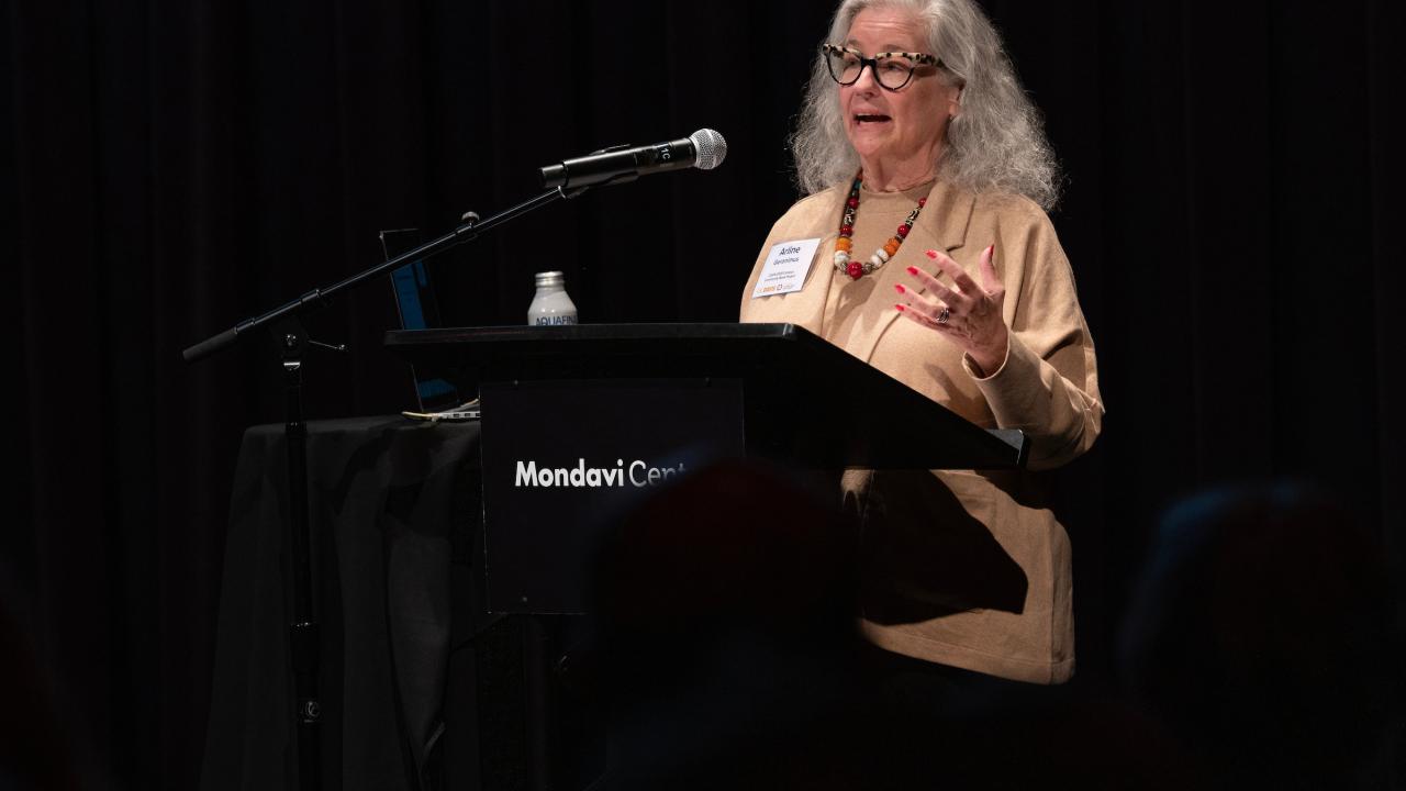 Standing a podium and illuminated by a stage light, Dr. Arline T. Geronimus speaks while staring at notes. She has long blonde hair past her shoulders, a white blouse and glasses with a red frame. 