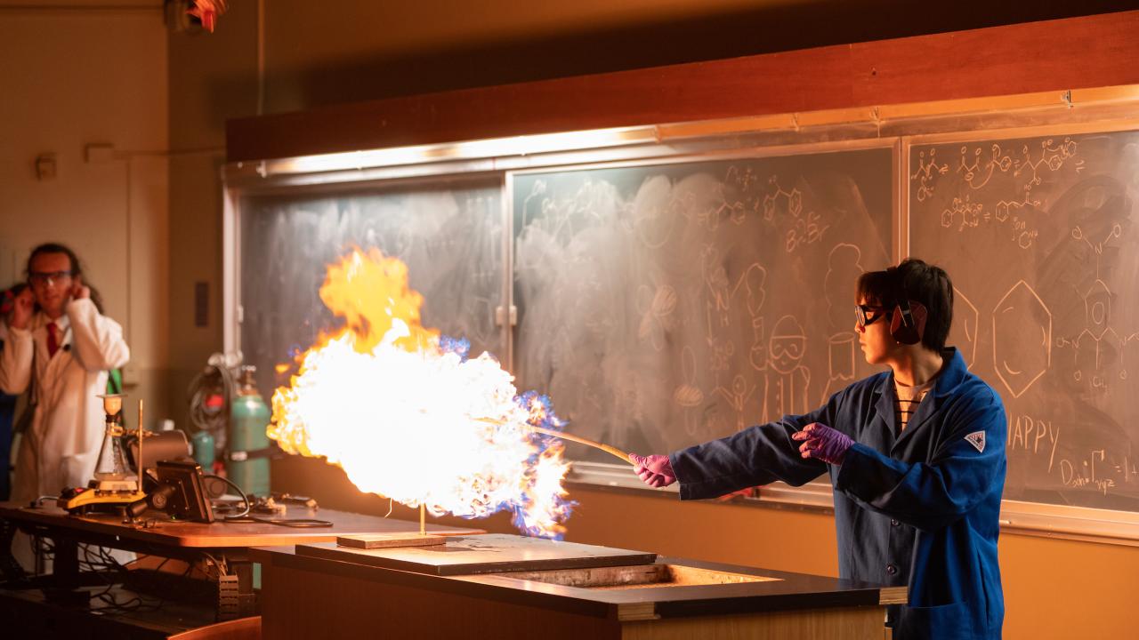 Inside a lecture hall, towards the right of the frame, a student in a lab coat with goggles on holds a stick that is engulfed in flames. The flames are caused by the demonstration the student is leading to a crowd at Rock Hall on UC Davis. On the left, a man in a lab coat plugs his ears at the sound of the controlled explosion. 