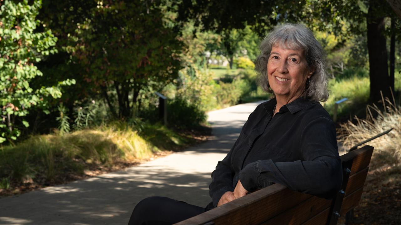 In an outdoor setting, Kate Scow sits on a bench towards the right of the photo, facing the camera and smiling. She's wearing a dark blue long sleeve top and is angled just slightly toward the camera. 