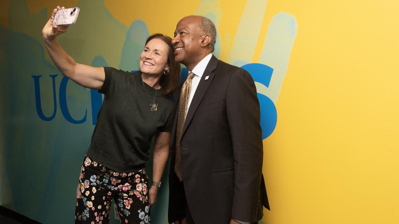A woman and a man smiling together while taking a selfie against a colorful background.