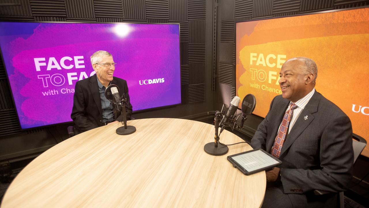 Two men sitting across from each other at a round table, microphones in front of them.