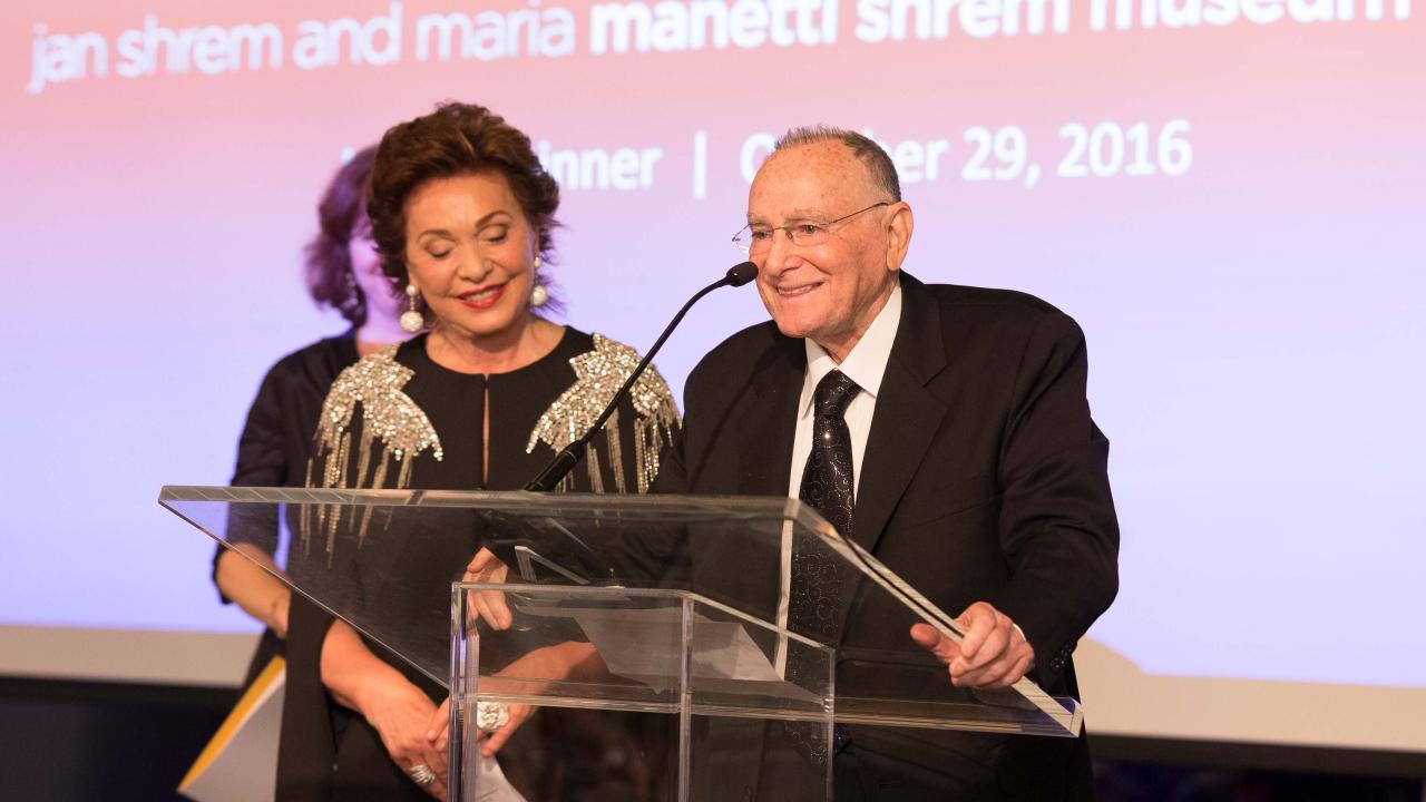 Woman and man in evening attire, at microphone, in front of backdrop of Manetti Shrem Museum sign in rainbow colors