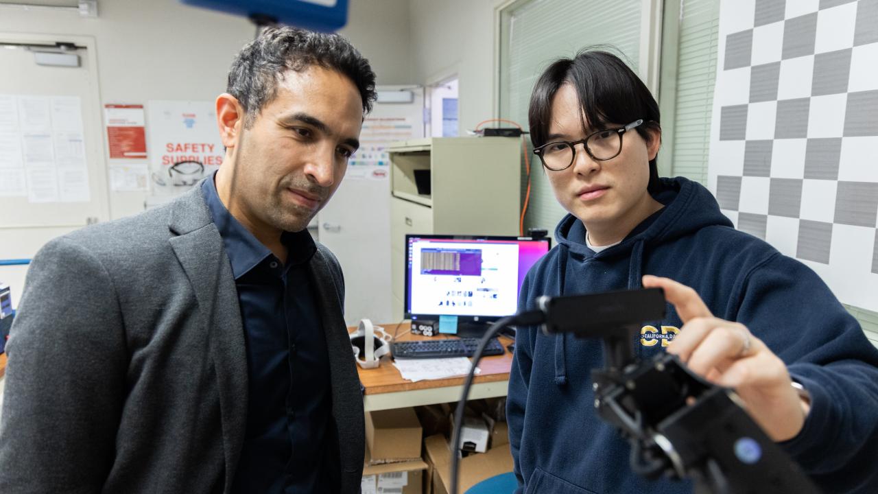 At left, a man with short curly dark hair wearing a grey jacket looks at a device held up by a younger man wearing a dark blue hoodie and glasses. 
