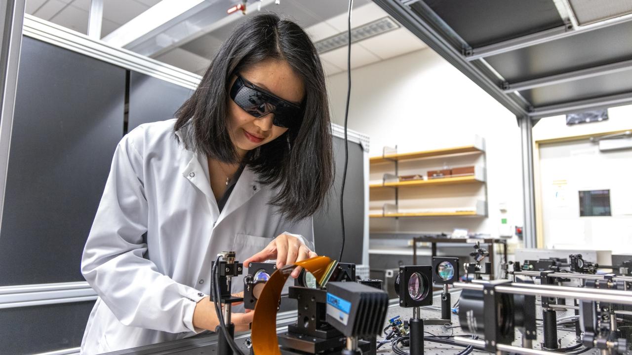A woman with dark hair wearing a white lab coat and dark wraparound eyeglasses leans over technical equipment on a lab workbench. 