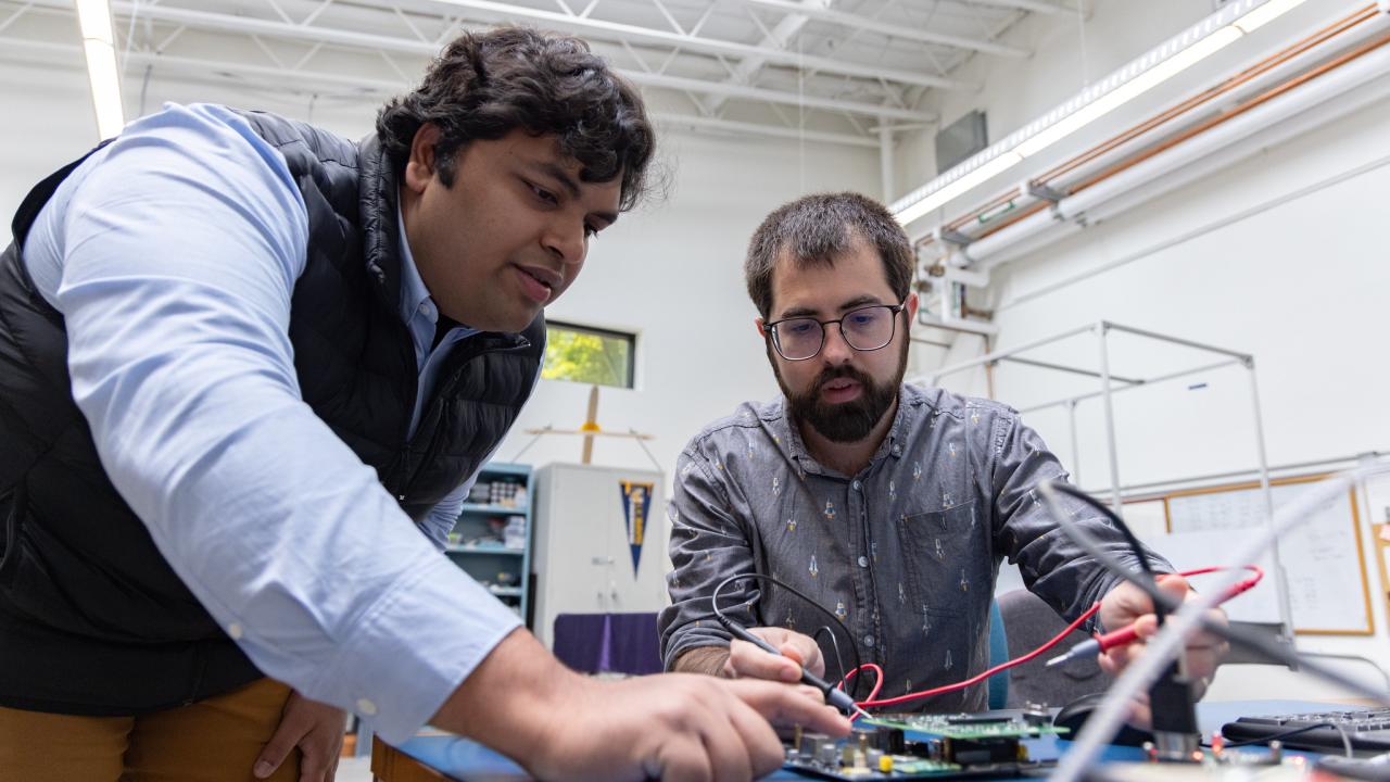 Two young men lean over a work table. Person on right, with short beard and glasses, is using a tool to test a circuit board as the person on left, with dark hair wearing a shirt and sleeveless vest, leans to examine it. 