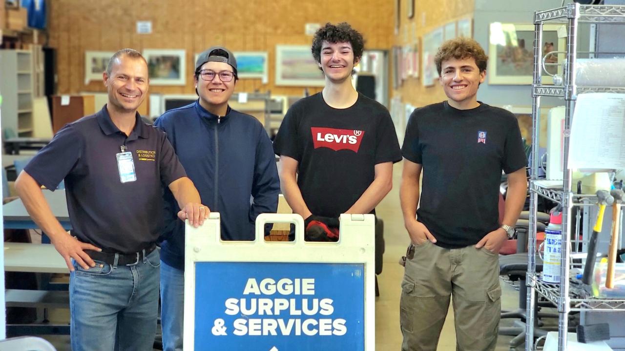 From left to right, AggieSurplus Coordinator Robert Juhler and Student Assistants Nick Gomez, Isaac Ewing and Lucas Morace stand and smile behind a sign that says "Aggie Surplus and Services." 