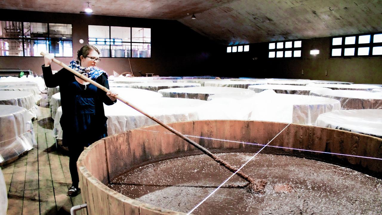 A person stirs something liquid in a big vat inside an industrial room.