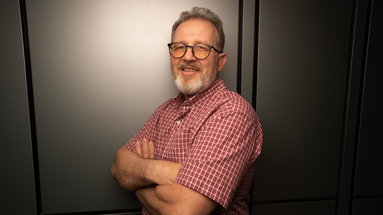 Professor Andreas Bäumler stands for a portrait with his arms crossed at a slight angle towards the camera, against a white backdrop inside an office setting. He is wearing a plaid red short sleeve shirt, glasses and has a full beard with hints of grey, and is smiling towards the camera. 