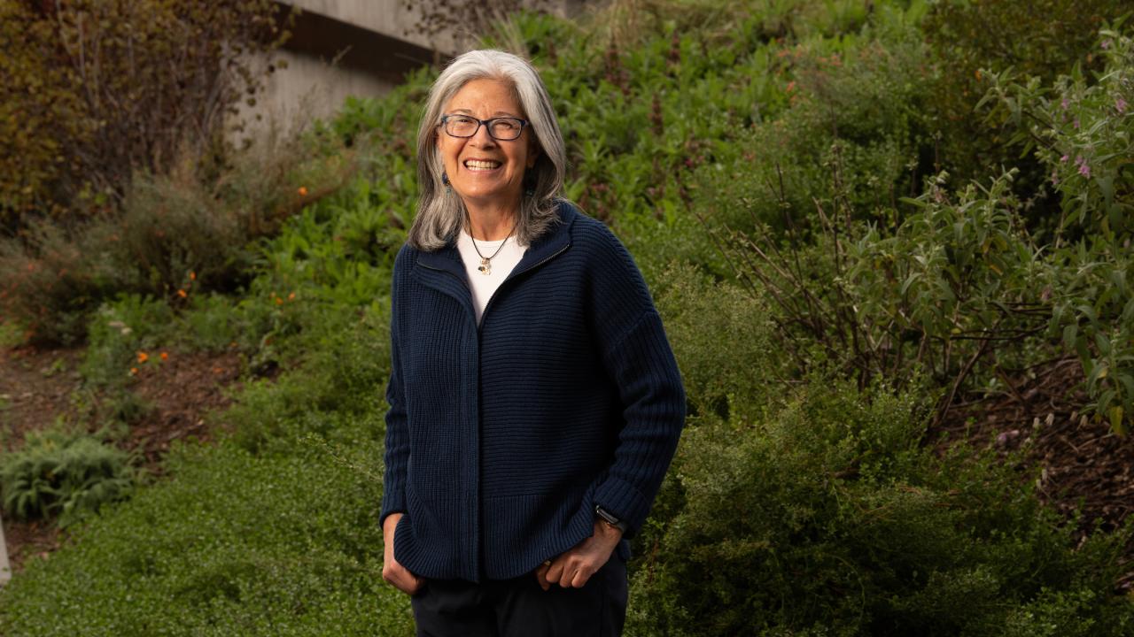 Outdoors in front of a grassy hill stands Professor Isabel Montañez, smiling towards the camera. She is wearing glasses, a blue button-up shirt with white undershirt and black pants. 