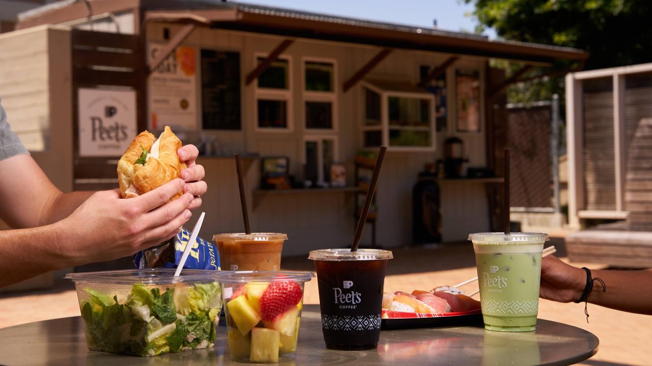 In an outdoor cafe setting, an unknown person is holding a sandwich above a table full of iced coffees, iced teas, salads and other food options. It's a sunny day with a blue sky peeking above the near food kiosk. 