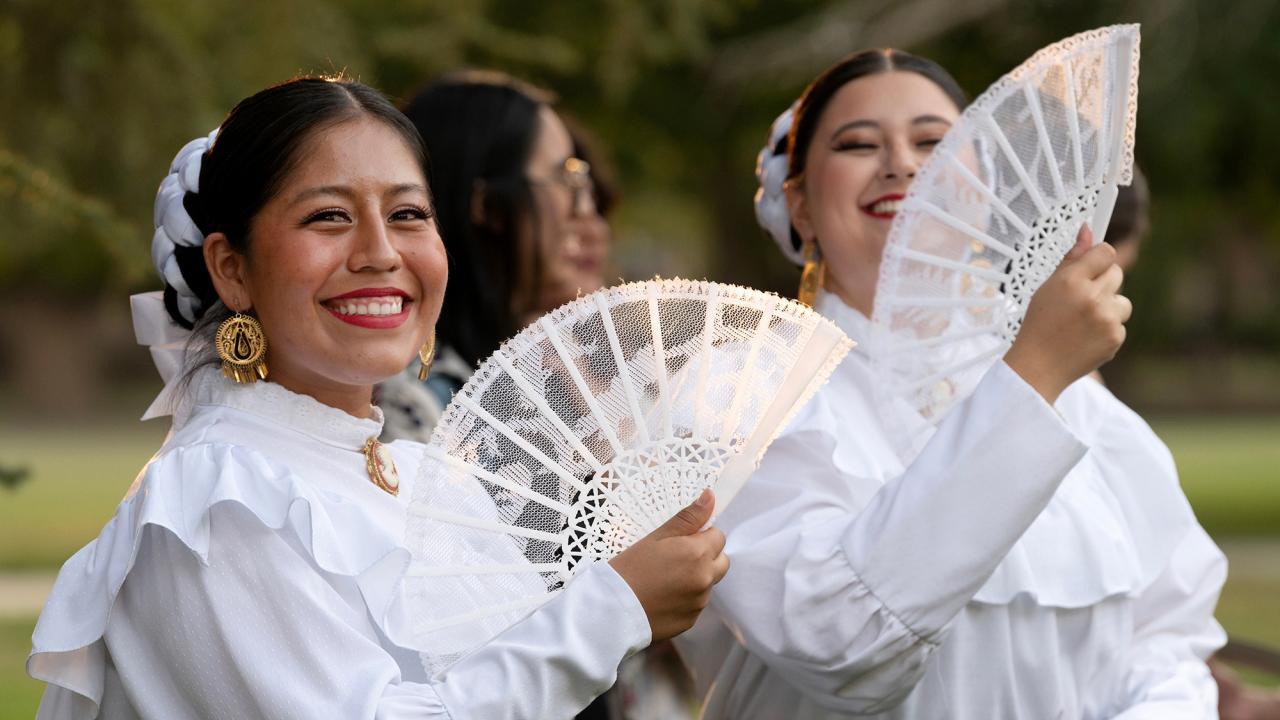 Two women smile while holding lace fans in a sunny outdoor setting.