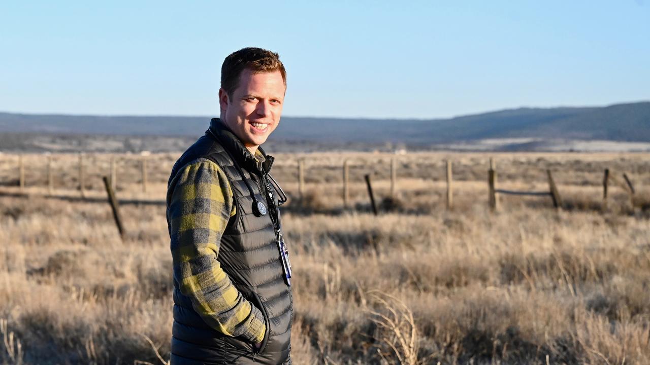 A man in a puffer vest and plaid short stands on a remote ranch, looking at the camera. Brushy fields and distant mountains are in the background.