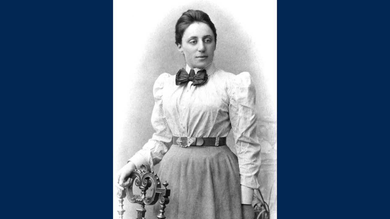 Black and white photo of a woman in early 20th century clothing with a white blouse, long skirt and bow at her throat. Her hand rests on the back of a chair. 