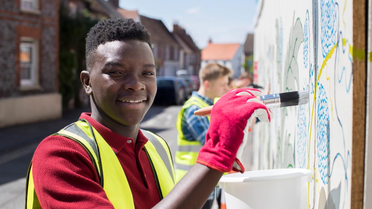 Black student wearing red holds paint brush near a wall mural with other teens in background