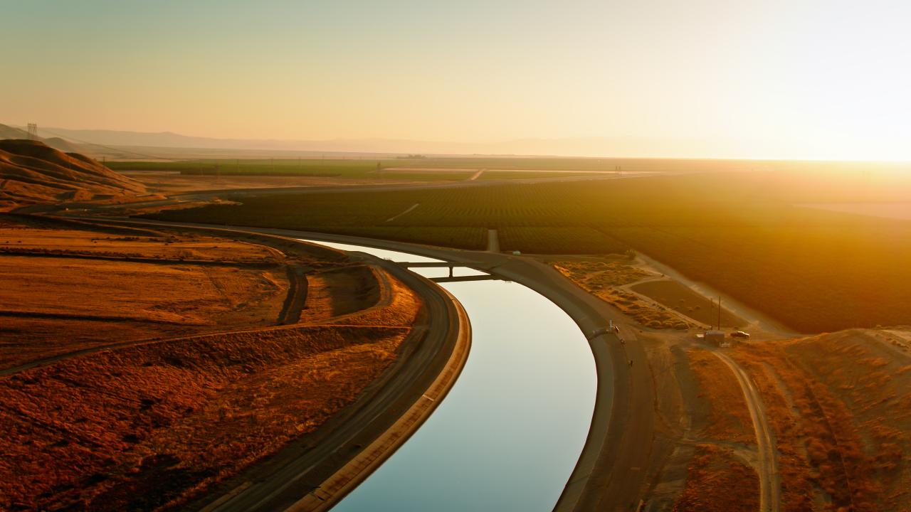Drone shot of California aqueduct curving between an auburn barren hillside and agricultural fields in Kern County at sunset