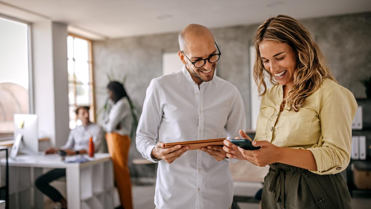 A man in a white shirt and black glasses and a woman in a yellow shirt with blonde hair look at the phone and tablet in their hands. In the background, a standing woman speaks to a man sitting at his desk in an office.