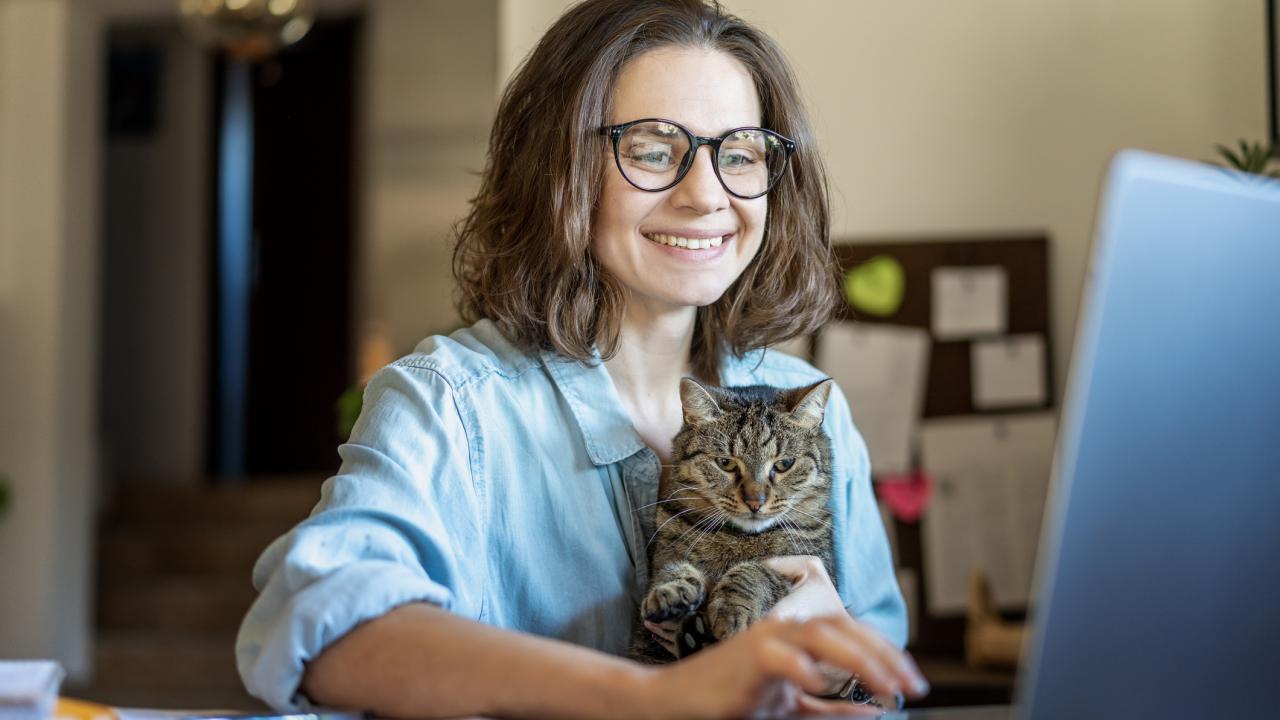 Woman using a laptop with a relaxes cat on her lap, representing how telehealth helps pet owers manage chronic conditions in cats from home. 