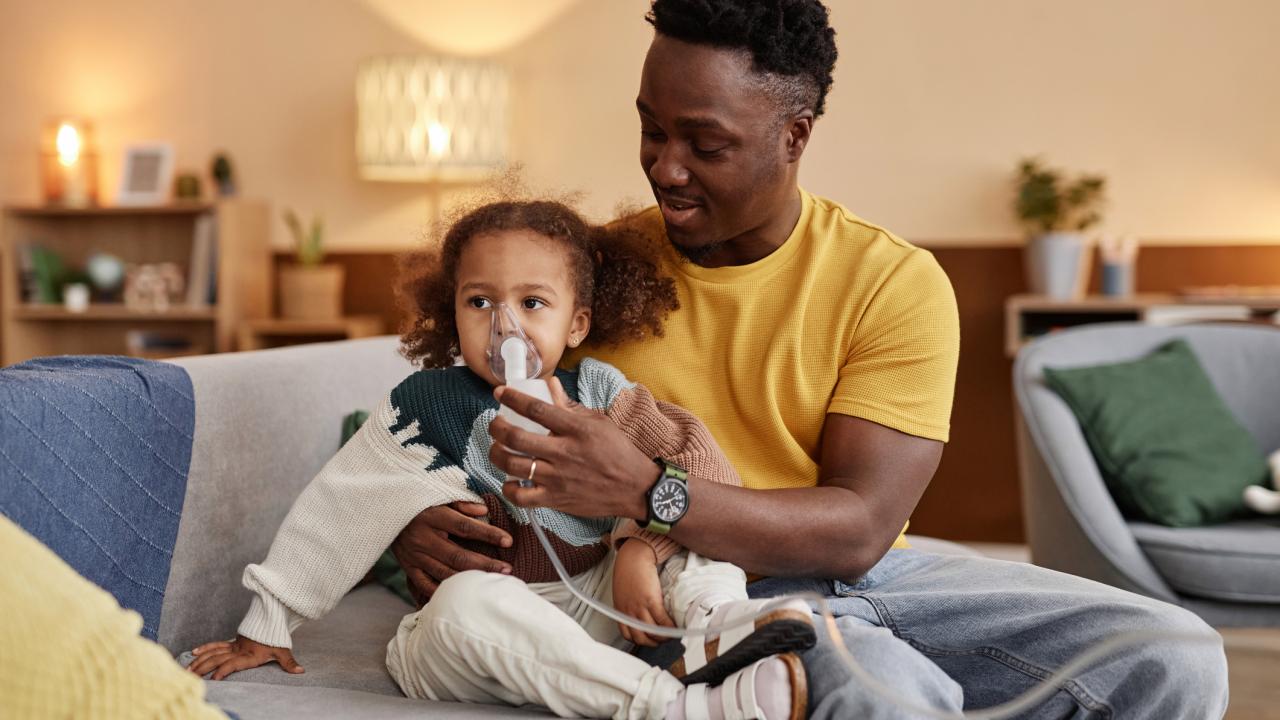 An African American man helps a child use an inhaler. They are sitting on a couch in a casual home setting. 