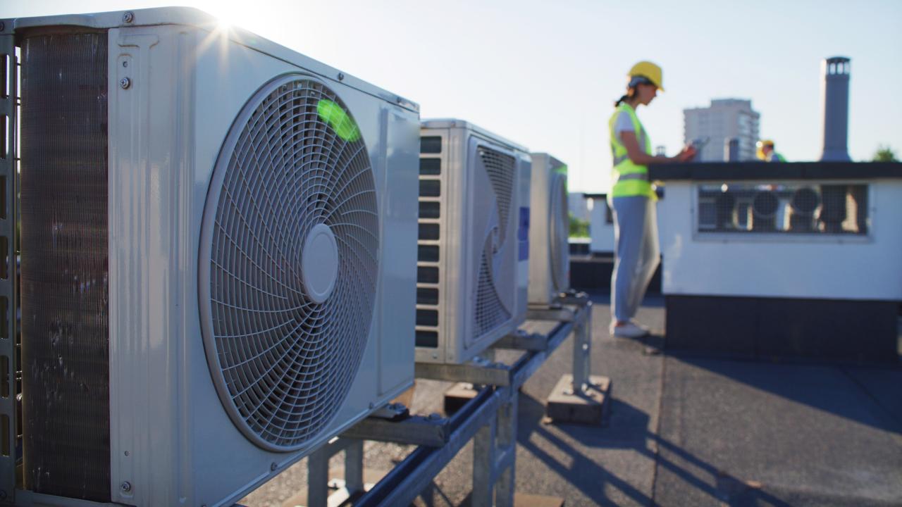Oblique view of air conditioner units on a rooftop. A technician wearing a high visibility vest and hard hat is in the background. 