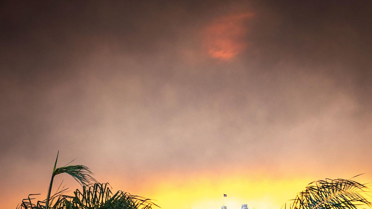 black and gray smoke plume moves over orange sunrise with golden light peaking below above palm trees and a house in Los Angeles
