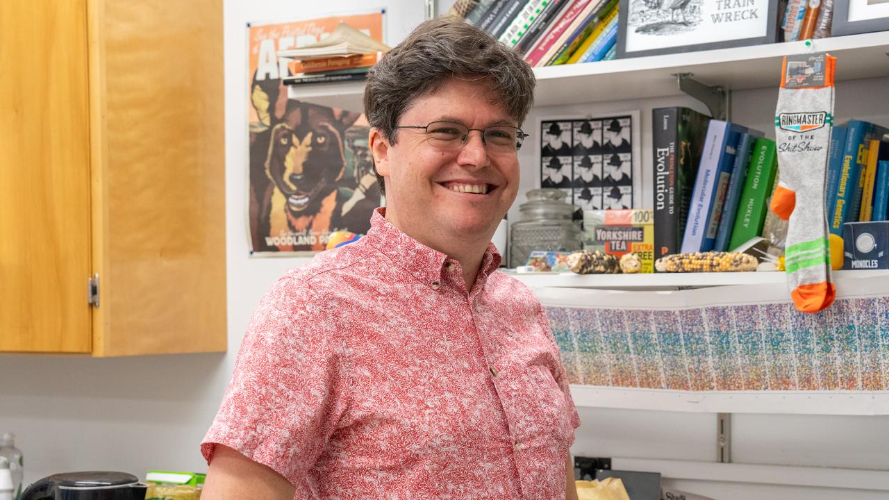 Head and upper body of a white man with short dark hair and glasses wearing a pink short sleeved shirt and smiling. Behind him are bookshelves. 