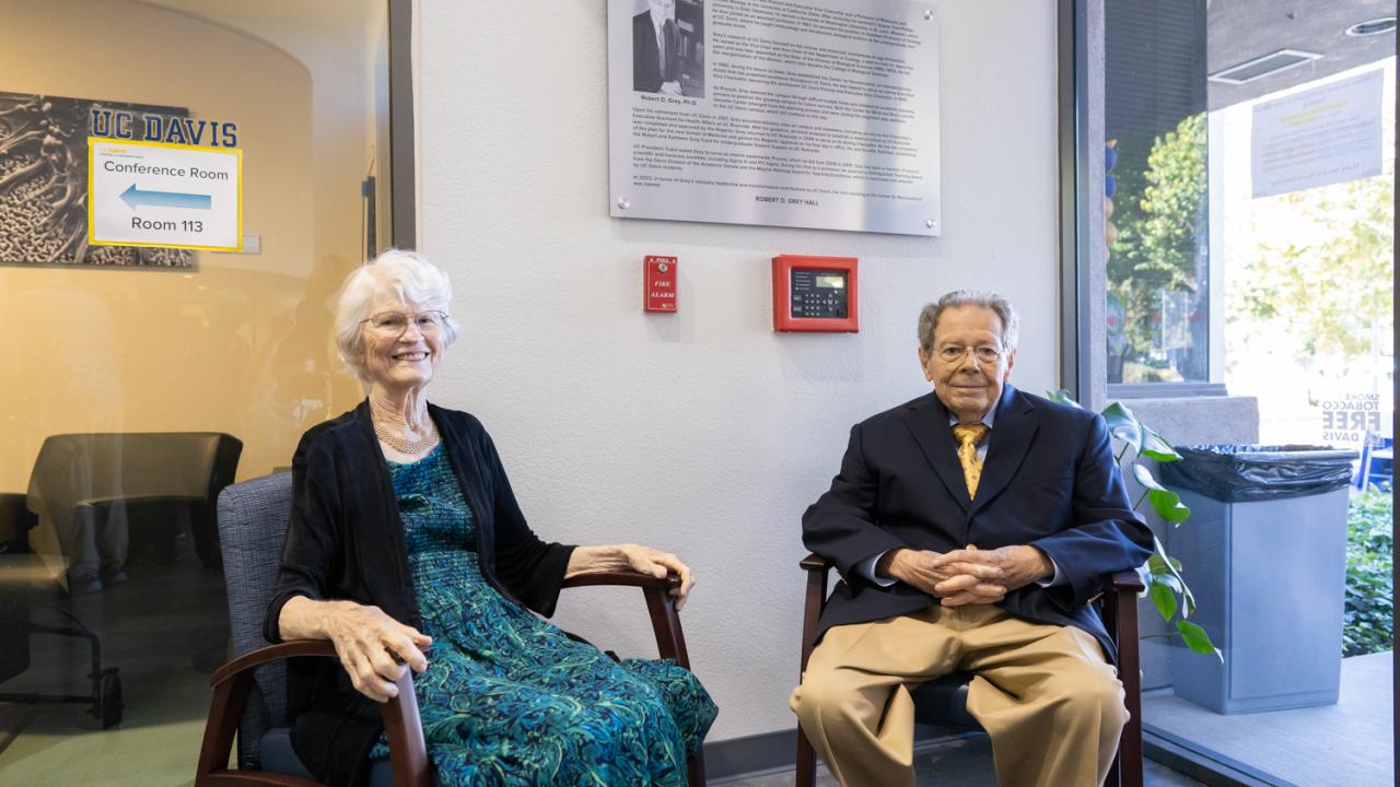 Elderly man and woman seated in a modern office space, smiling and dressed formally.