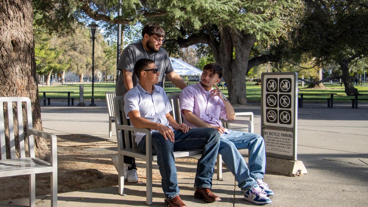 Three students chat and sit on benches in a shady spot near the UC Davis Quad. A “No Bicycle Parking” sign is visible nearby, along with tall trees and a white tent in the background.