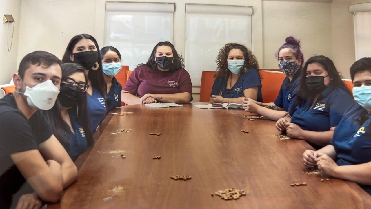 A group of masked UC Davis student staff and leaders sit around a large conference table inside the Native Nest. They smile behind their masks, dressed in matching navy UC Davis polos and casual clothes.