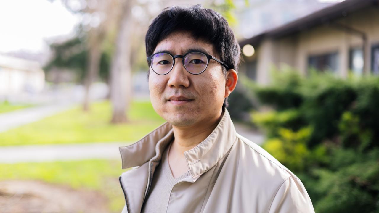 A man with glasses stands outdoors, wearing a light jacket, with greenery in the background.