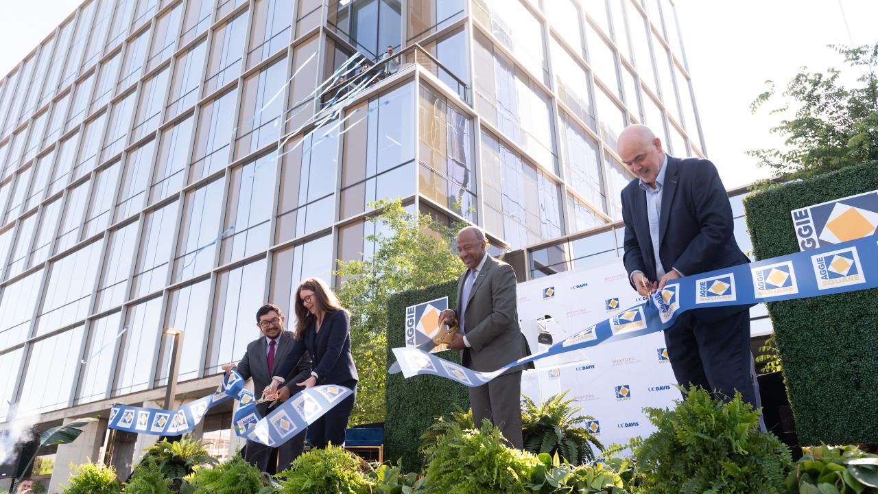 Outdoor setting, large glass building in background. Four people stand in a row cutting a ribbon into pieces. 