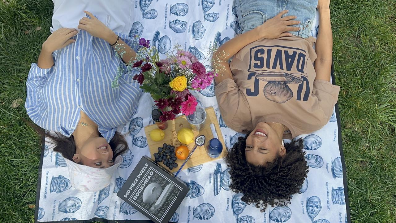 An upside-down view of two smiling female Aggies laying on the grass on an Egghead-designed blanket separated by fruit, flowers and several Egghead branded items including a jigsaw puzzle, cutting board and pen