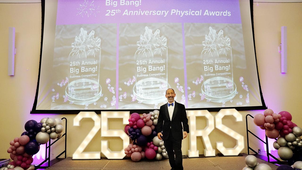 A man in a tuxedo stands on a stage festooned with grape-like orbs, a lit sign reading "25 YRS" and a banner reading, "Big Bang! 25th Anniversary Physical Awards"