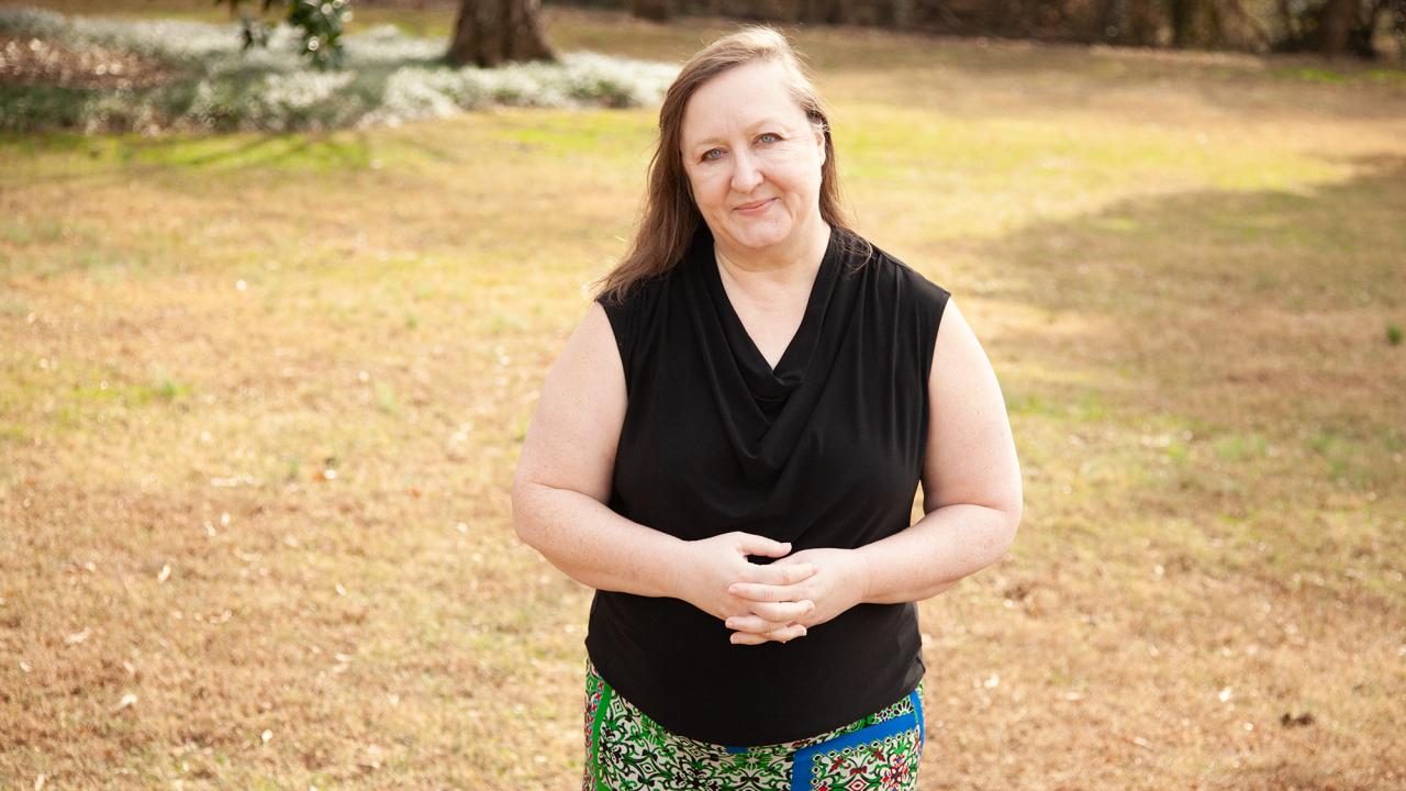 A woman with shoulder length blonde hair wearing a black sleeveless top and patterned skirt stands outdoors with her hands clasped in front. 