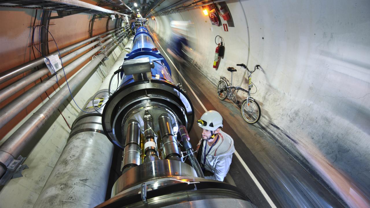 A worker in a helmet inspects large metallic pipes inside a tunnel.