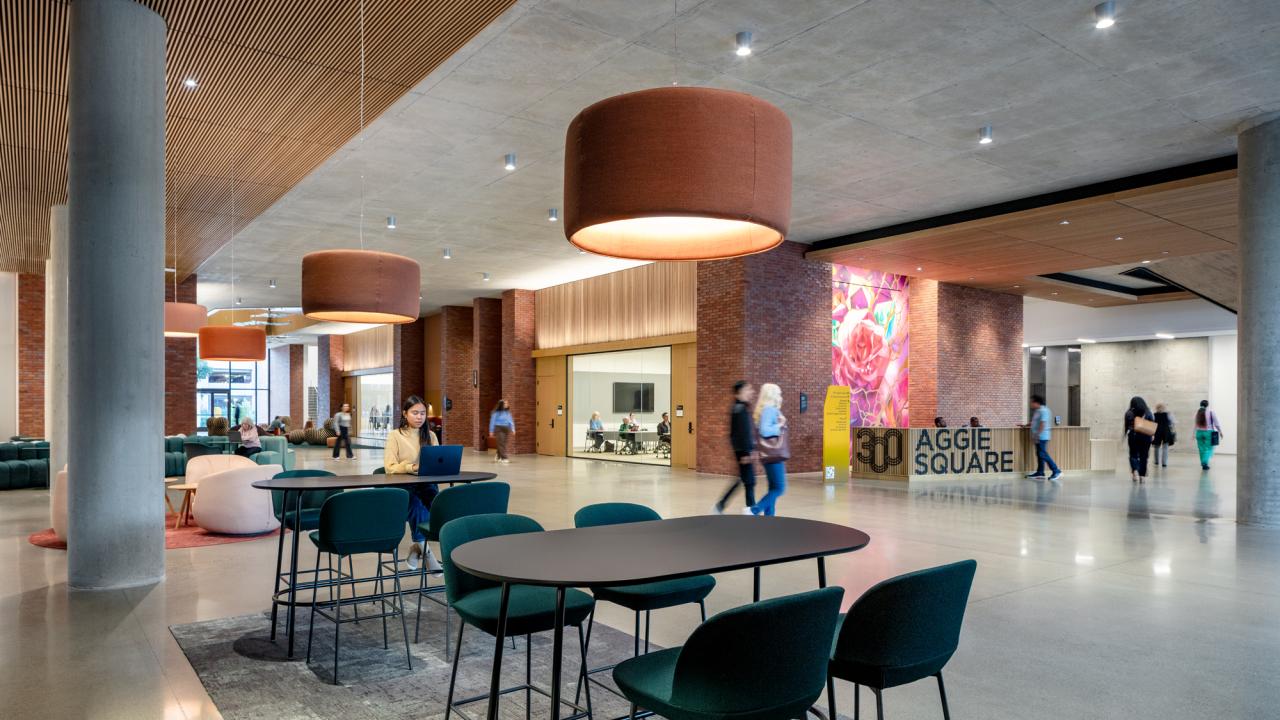 Modern lobby with light fixtures, tables and seating areas, and people interacting. A reception desk has a "300 Aggie Square" banner on it.