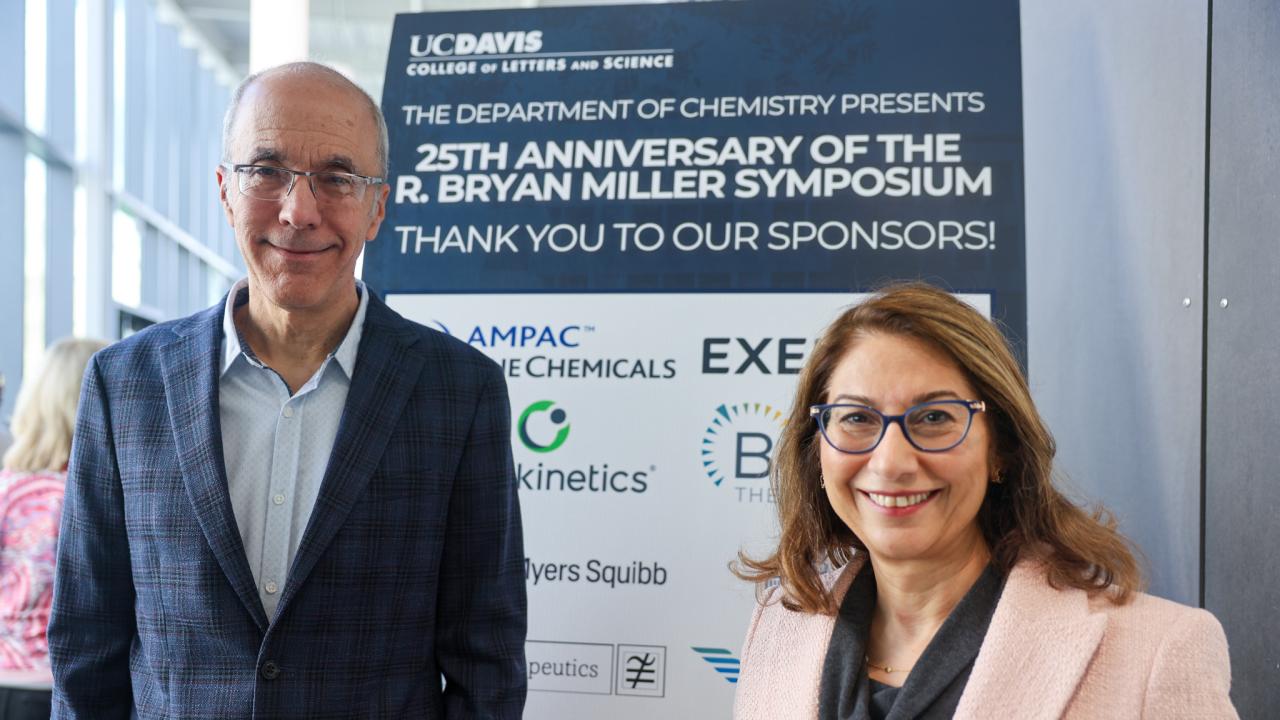 Left, a man with cropped hair and glasses wearing a grey jacket; right, a shorter woman with shoulder length brown hair, glasses and a pink jacket. They stand in front of a poster for the 25th Anniversary of the Bryan Miller Symposium with a list of sponsors. 
