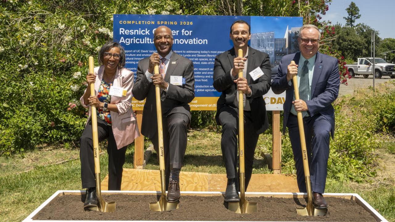 Five people in business attire holding gold shovels at an outdoor groundbreaking