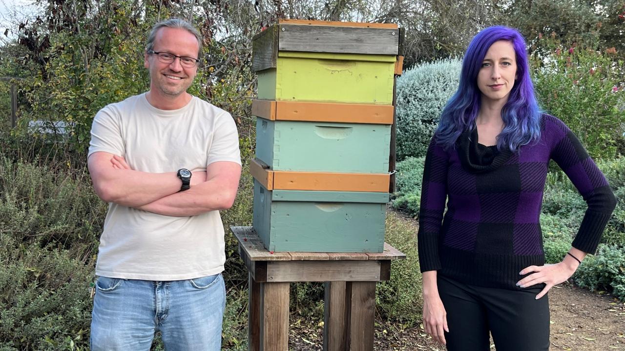Sascha Nicklish in ponytail, white t-shirt and jeans stands with Julia Fine in black shirt and pants and purple hair stand on either side of a wooden, mint green and yellow bee hive. 