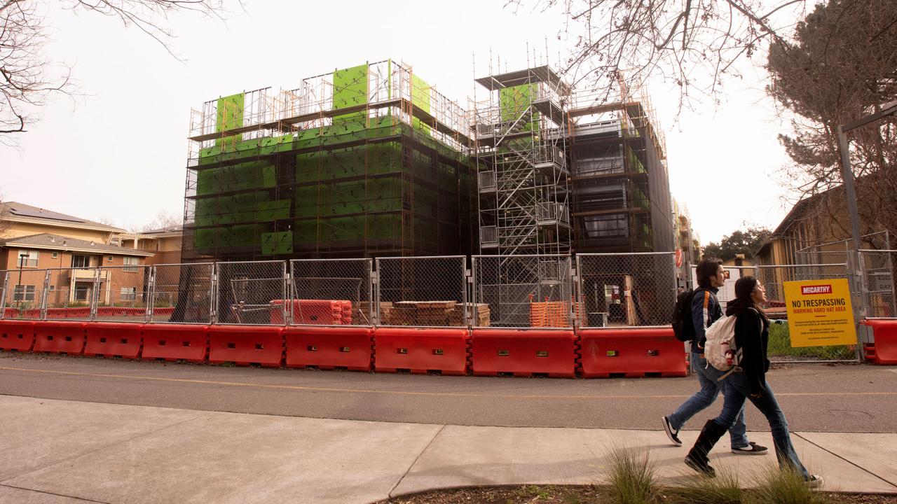 Two students walk past a construction site with scaffolding and safety barriers.