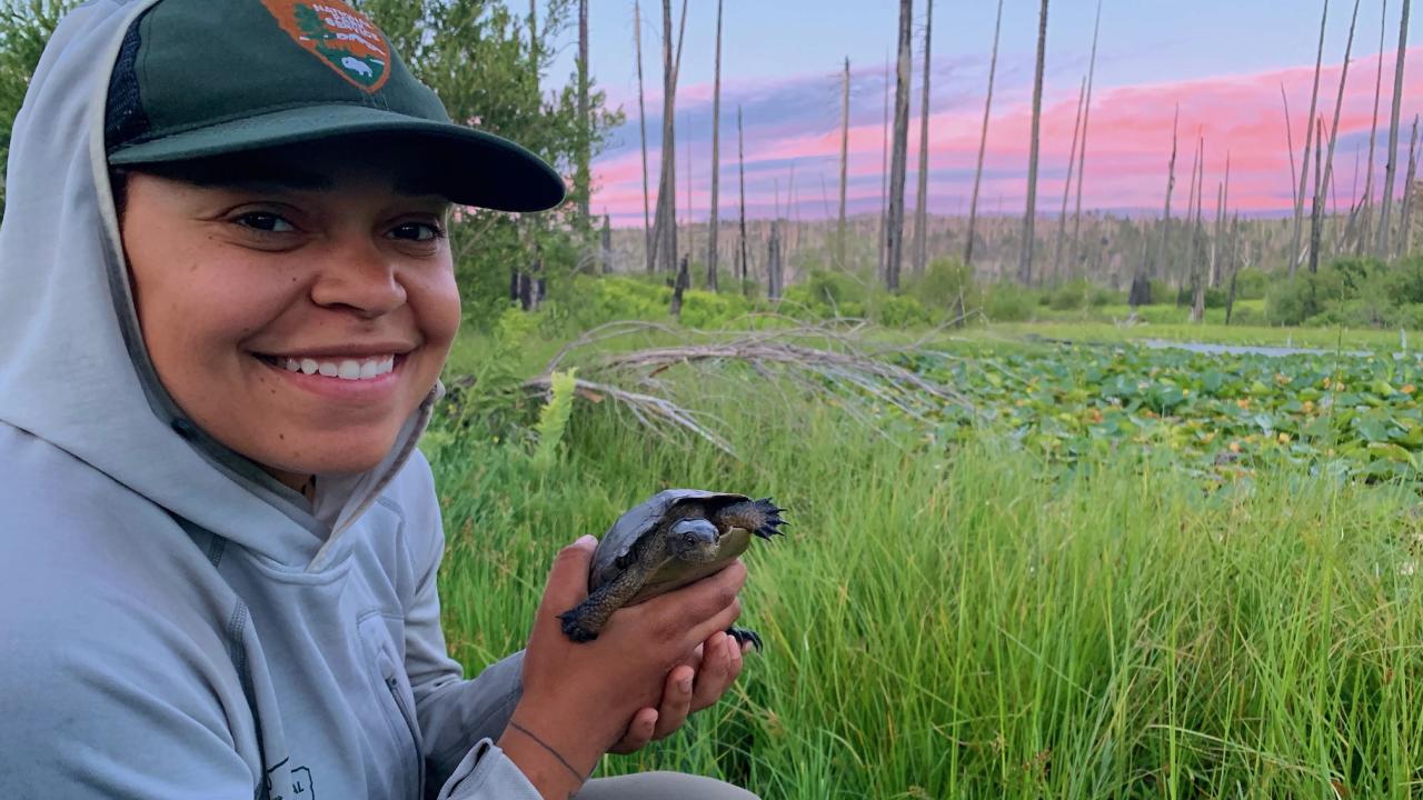 Sidney Woodruff in gray hoodie and NPS ballcap holds northwestern pond turtle while crouching beside a pond with pink sunset in background