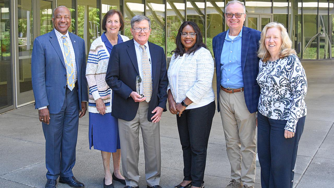 A group of six professional dressed men and women stand outside a venue, smiling and staring at the camera. The group includes 