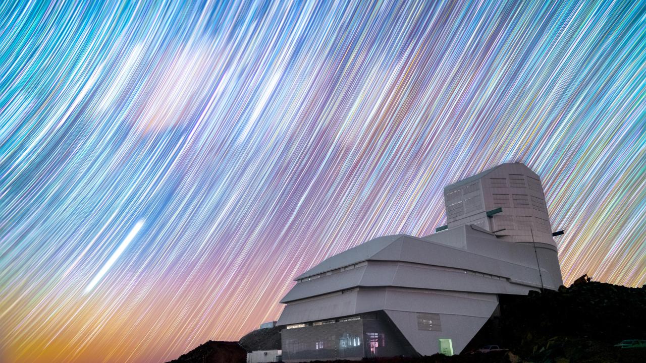 Foreground, a futuristic building on a rocky foundation. Background, brightly colored lines curve across a blue sky.