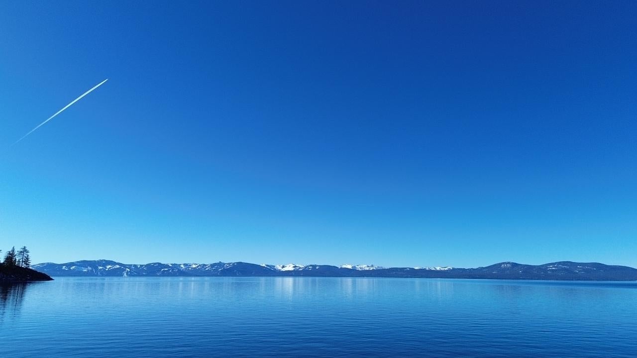 A plane's contrails streaks through a blue sky over deep blue Lake Tahoe